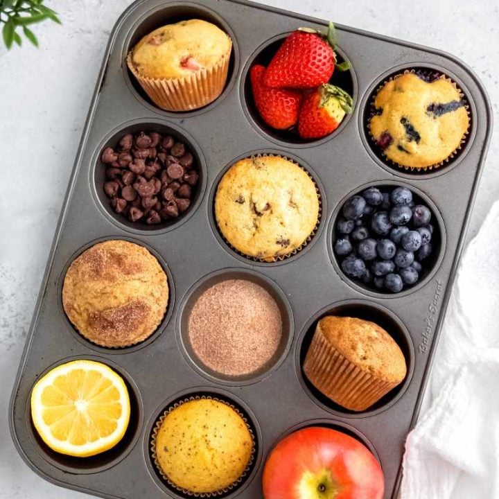 A muffin tin filled in alternating cups with six flavors of muffins and their corresponding mix-ins, such as strawberries, chocolate chips, blueberries, and more.