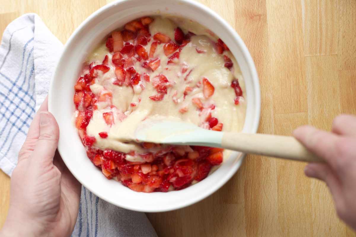 Hands use a wooden spatula to stir chopped berries into a batter