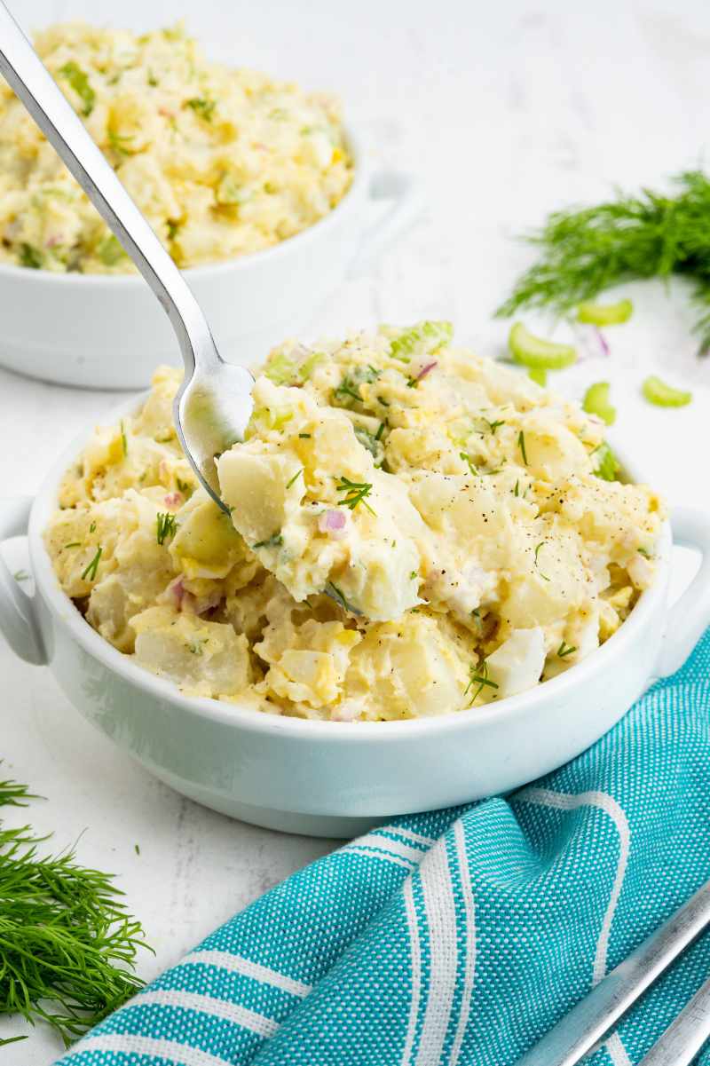 A fork lifts a bite of creamy potatoes from a small white serving bowl beside a teal and white linen napkin.