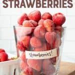 Frozen strawberries piled high in a labeled glass jar resting on a wooden cutting board. A text overlay reads, "How to Freeze Strawberries."