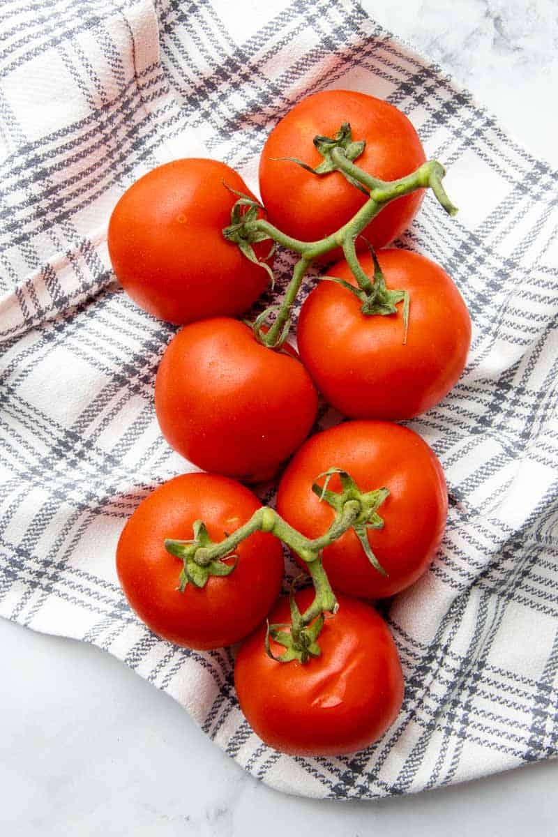 Overhead of seven fresh slicing tomatoes still on the vine sitting atop a kitchen towel.