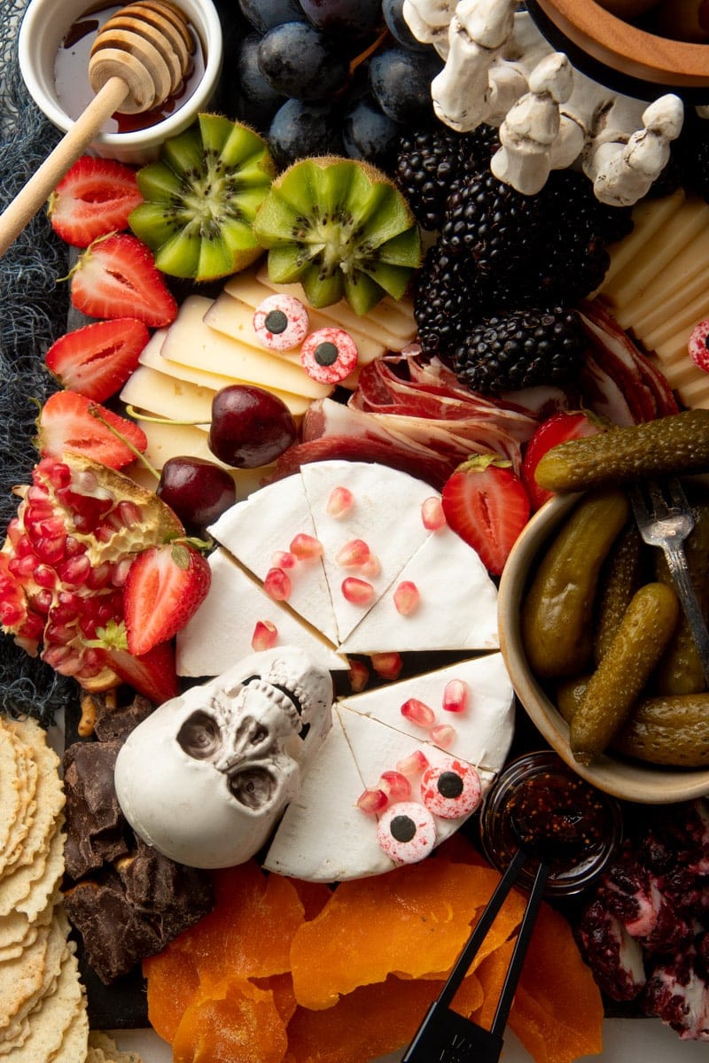 A decorative skull rests on a wheel of brie topped with pomegranate arils and surrounded by meats, cheeses, crackers, and fruits on a charcuterie board.