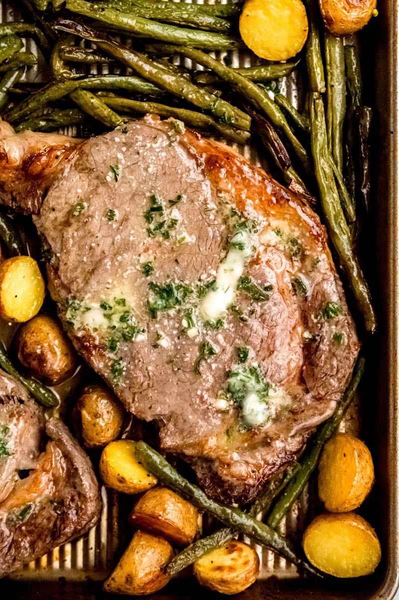 Close view of a garlic butter steak surrounded by veggies on a baking sheet.