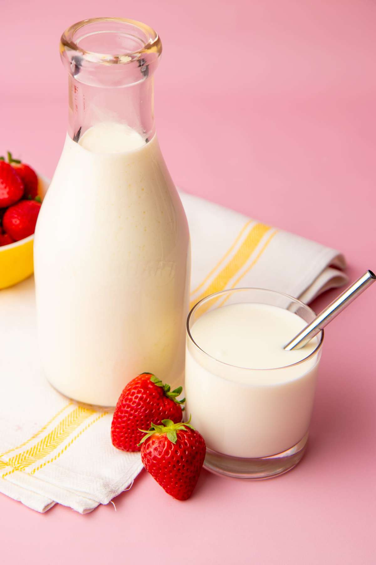 A full tumbler of homemade kefir with a metal straw sits with two fresh strawberries and a full glass carafe alongside it.