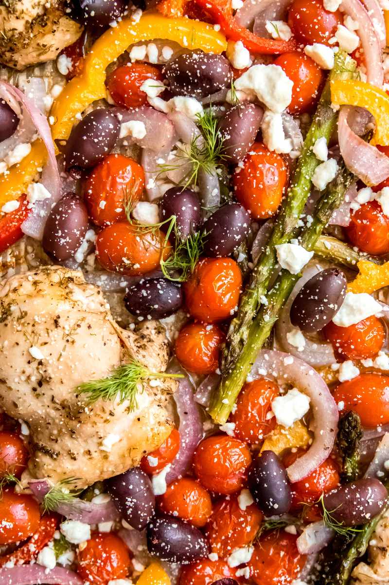 Close view of baked chicken thighs with cherry tomatoes, kalamata olives, bell peppers, asparagus, and onions all tossed in greek herbs and feta cheese.