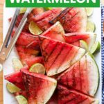 A rectangular white plate piled high with grilled watermelon slices garnished with lime wedges and tajin seasoning. A text overlay reads, "Grilled Watermelon."