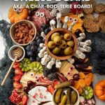 Overhead of a Halloween charcuterie board on a slate platter filled with meats, cheeses, fruits, nuts, crackers and spooky decorations. A text overlay reads, "Spooky! Halloween Cheese Board. AKA: A Char-Boo-Terie Board!"