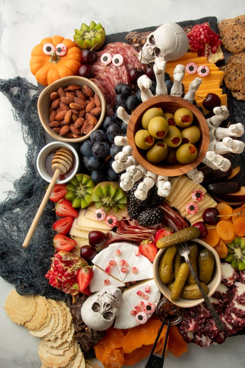 Halloween decorations adorn a spooky charcuterie board filled with cured meats, cheeses, fruits, and crackers on a black slate serving platter.