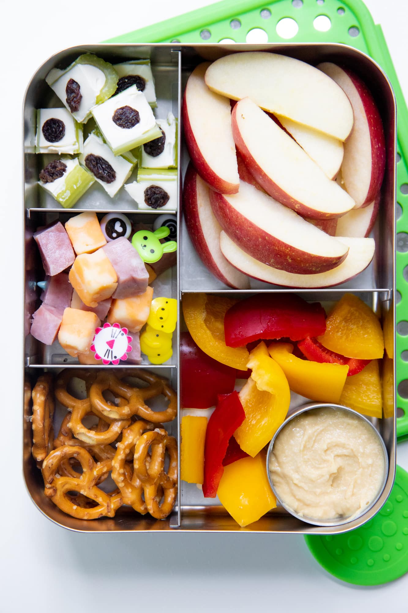 Close-up of a bento-style stainless steel lunch box filled with apple slices, pretzels, ham and cheese on toothpicks, bell peppers and hummus, and stuffed celery.