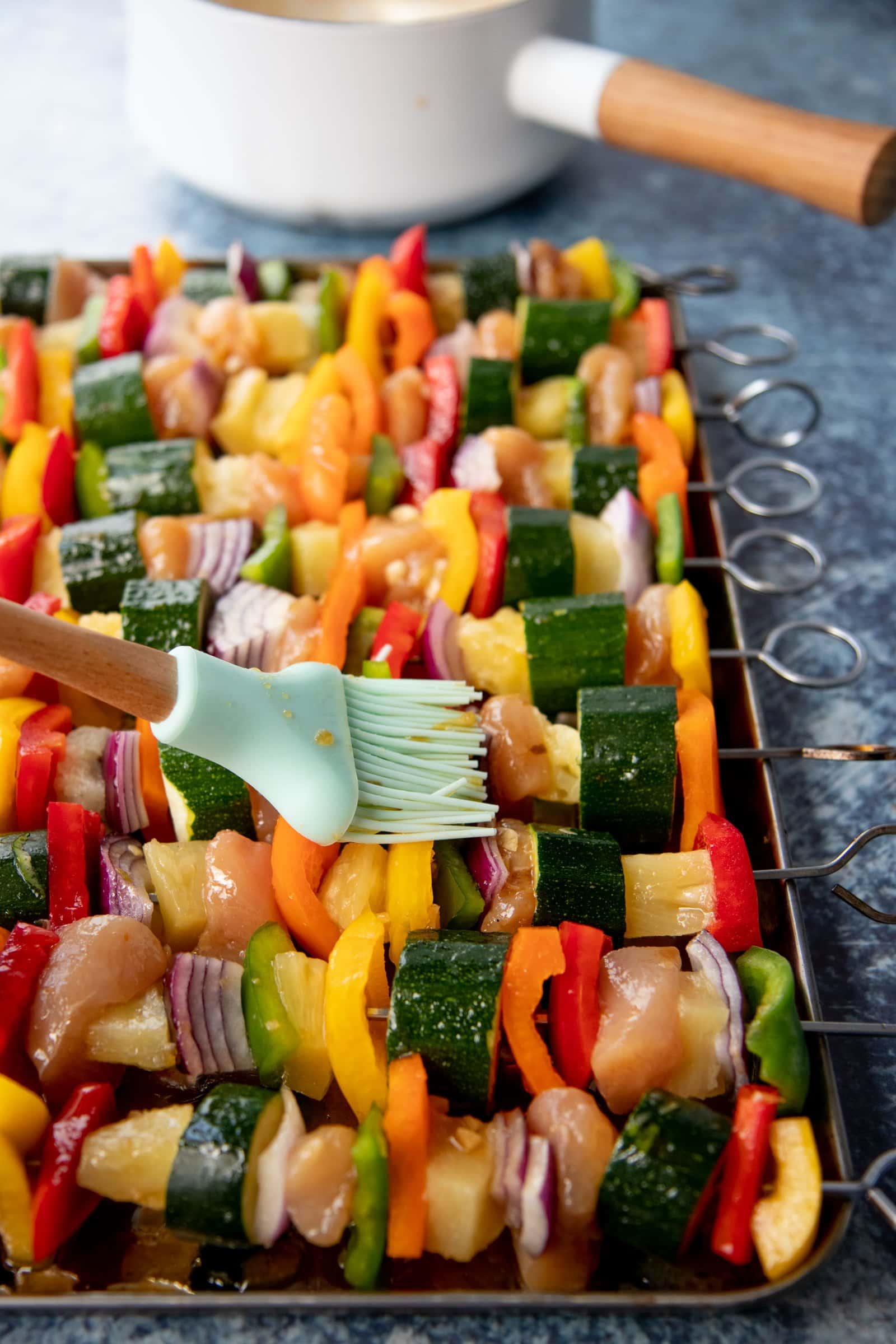 Pastry brush brushing marinade onto a tray of metal skewers threaded with chicken, vegetables, and pineapple to make Grilled Chicken Kabobs