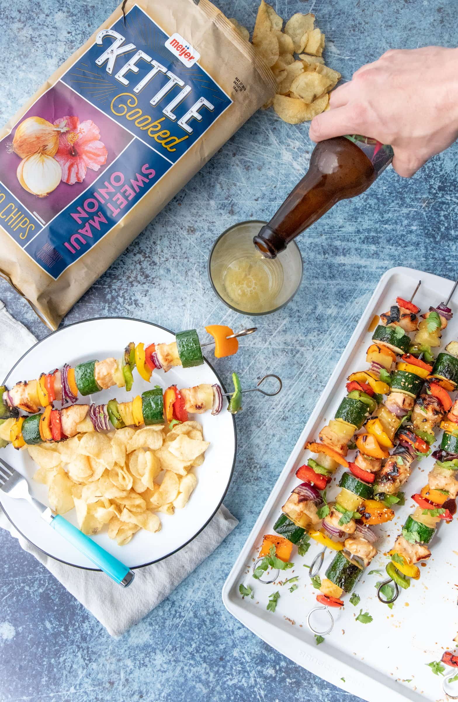 Hand pouring beer into a glass on a table full of Grilled Hawaiian Chicken Kabobs and kettle chips