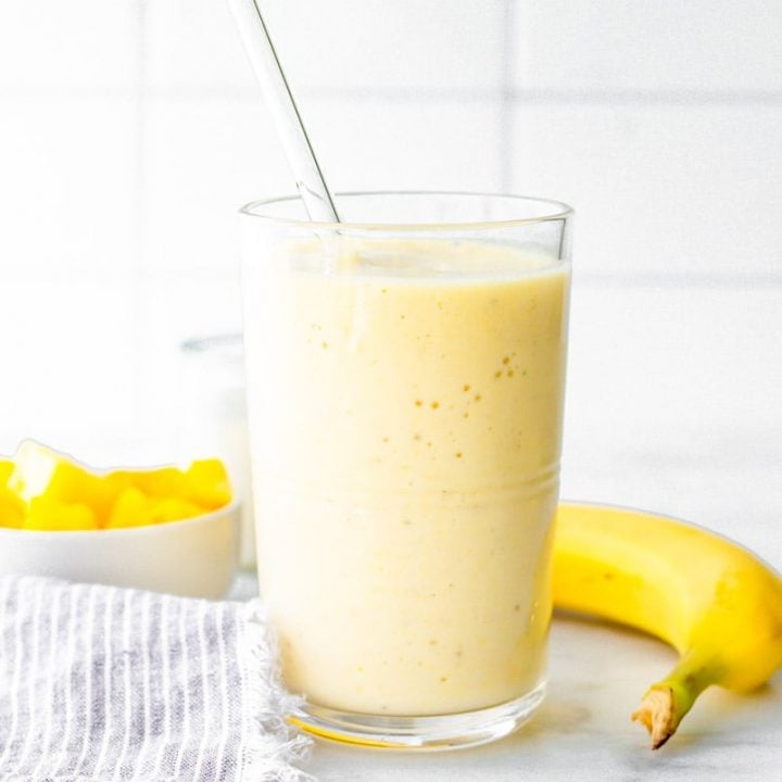 Coconut pineapple smoothie with a glass straw on a counter with fresh pineapple and banana.