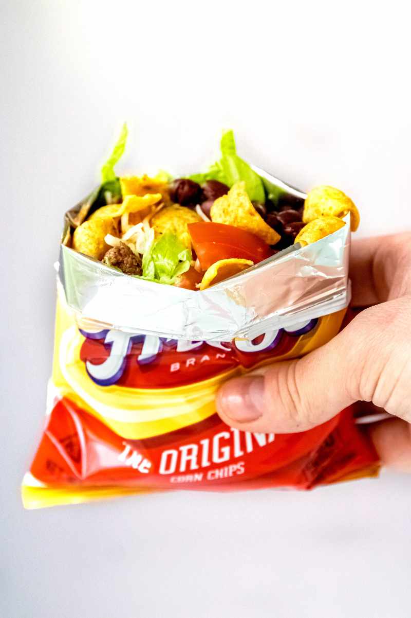 A hand holds up a walking taco made in a bag of corn chips on a light background.