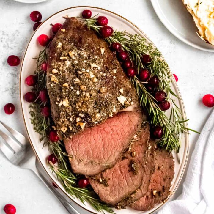 Top view of a holiday roast topped with herbs and garlic on an oval platter with fresh rosemary and cranberries around it.