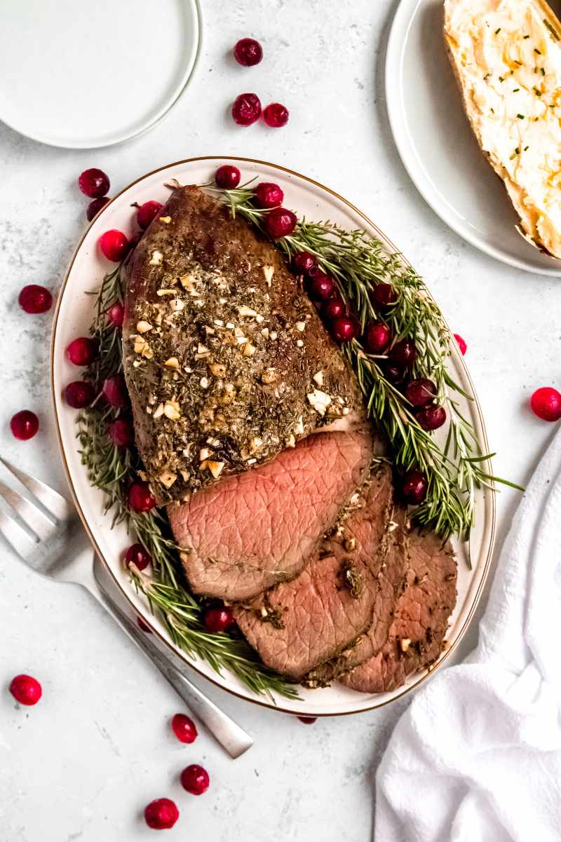 Top view of a holiday roast topped with herbs and garlic on an oval platter with fresh rosemary and cranberries around it.