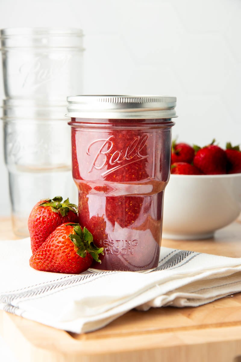 A full jar of strawberry jam sits on a folded tea towel in front of a bowl of strawberris.