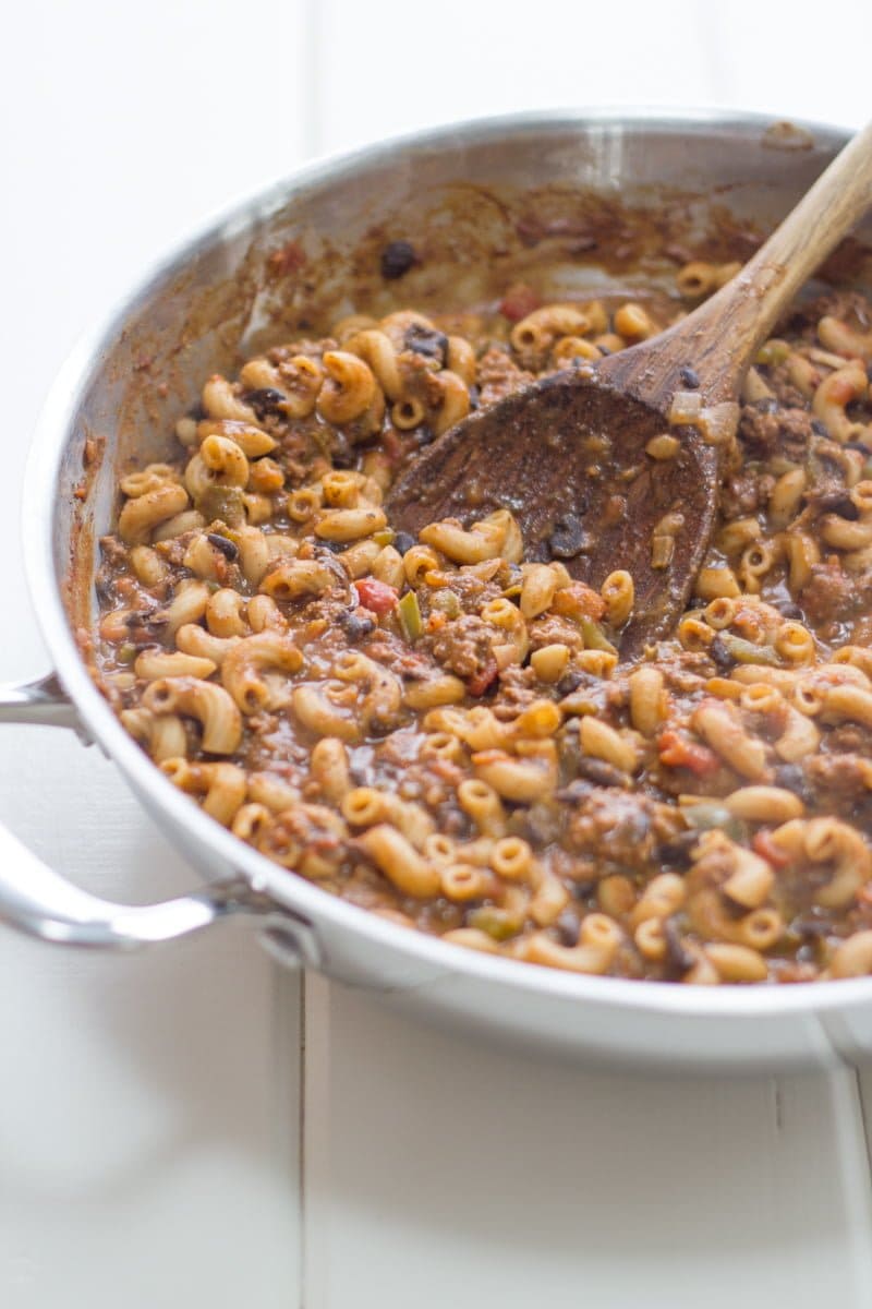 A pot of pasta and beef sits in a silver pan and is stirred by a wooden spoon.