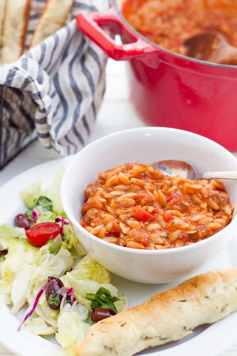 A bowl of Pasta e Faglioli sits on a white plate, surrounded by lettuce and a breadstick. A red dutch oven is seen in the background.