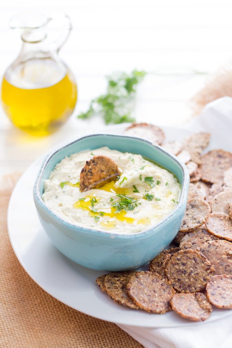 White bean dip drizzled with olive oil is in a light blue bowl, surrounded by crackers on a white platter.