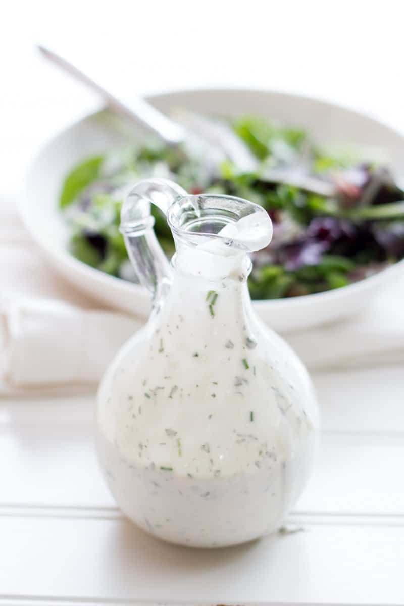 Glass bottle filled with healthy ranch dressing sits in front of a white bowl filled with salad.