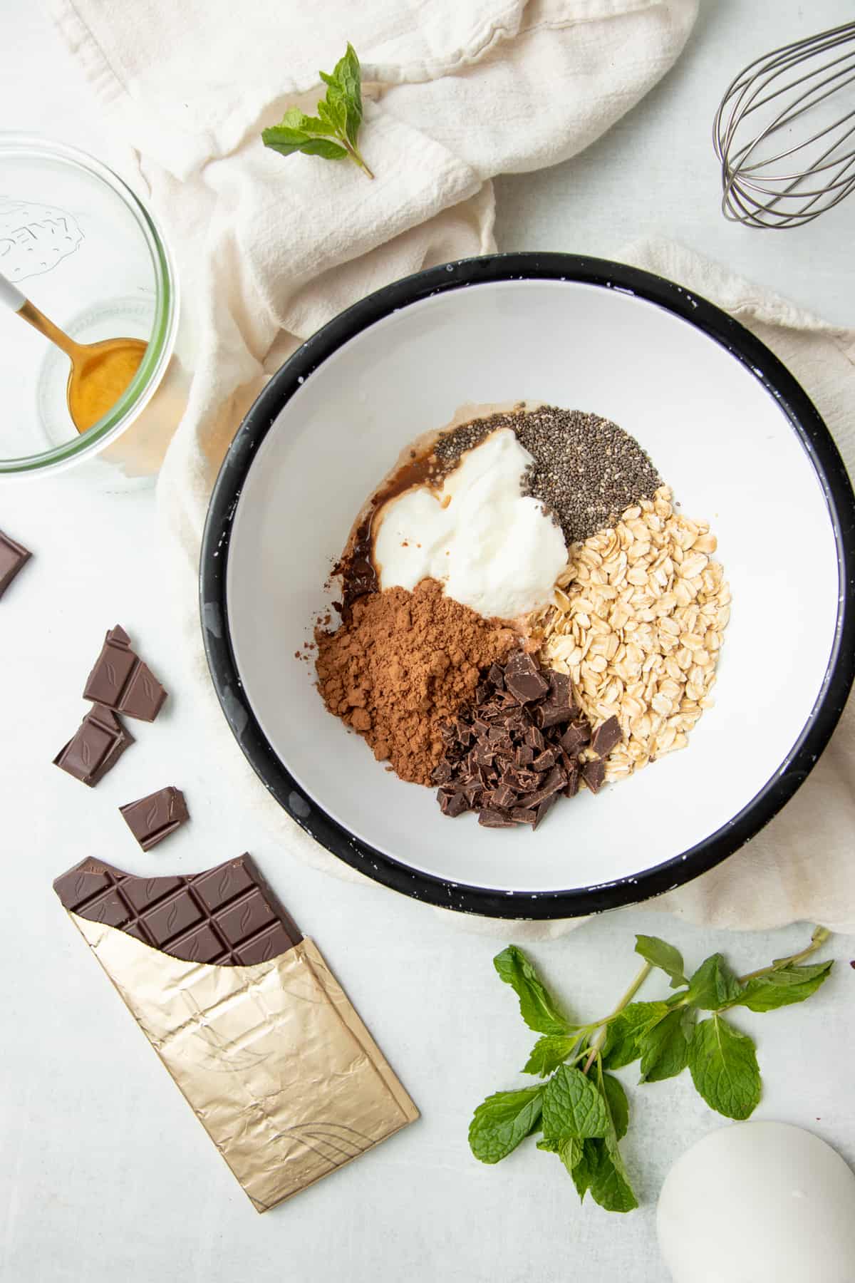 Overnight oat ingredients are arranged in a large white bowl with a black rim. A gold spoon in a glass jar sits to the side.