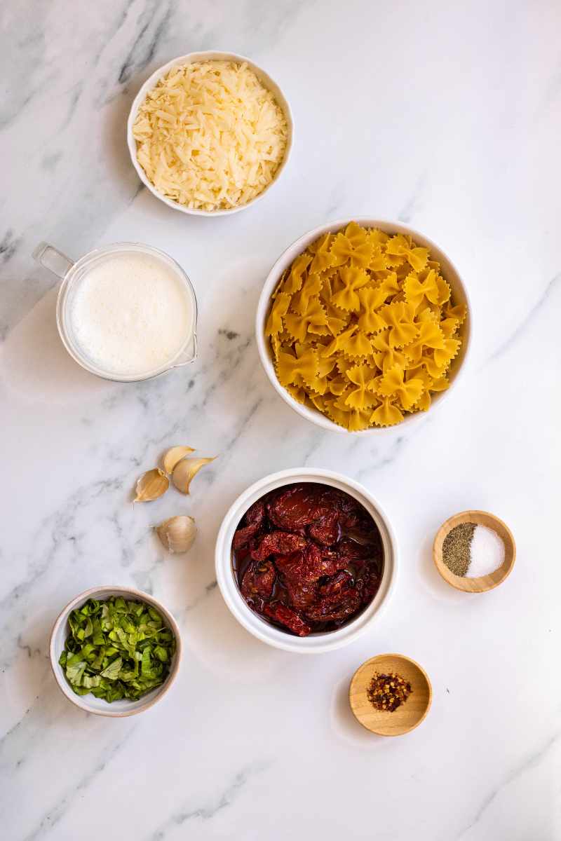 Overhead of ingredients for a pasta dish, including bowtie pasta, garlic cloves, fresh basil, and red pepper flakes