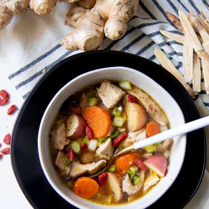 A spoon with a white handle sits in a white bowl full of herbal chicken soup. A knob of ginger is behind the bowl.