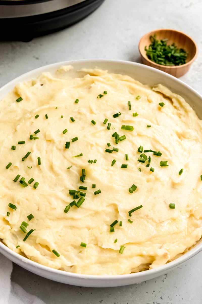 A wide serving bowl of Instant Pot mashed potatoes garnished with chives rests on a light grey counter with a small bowl of chives nearby.