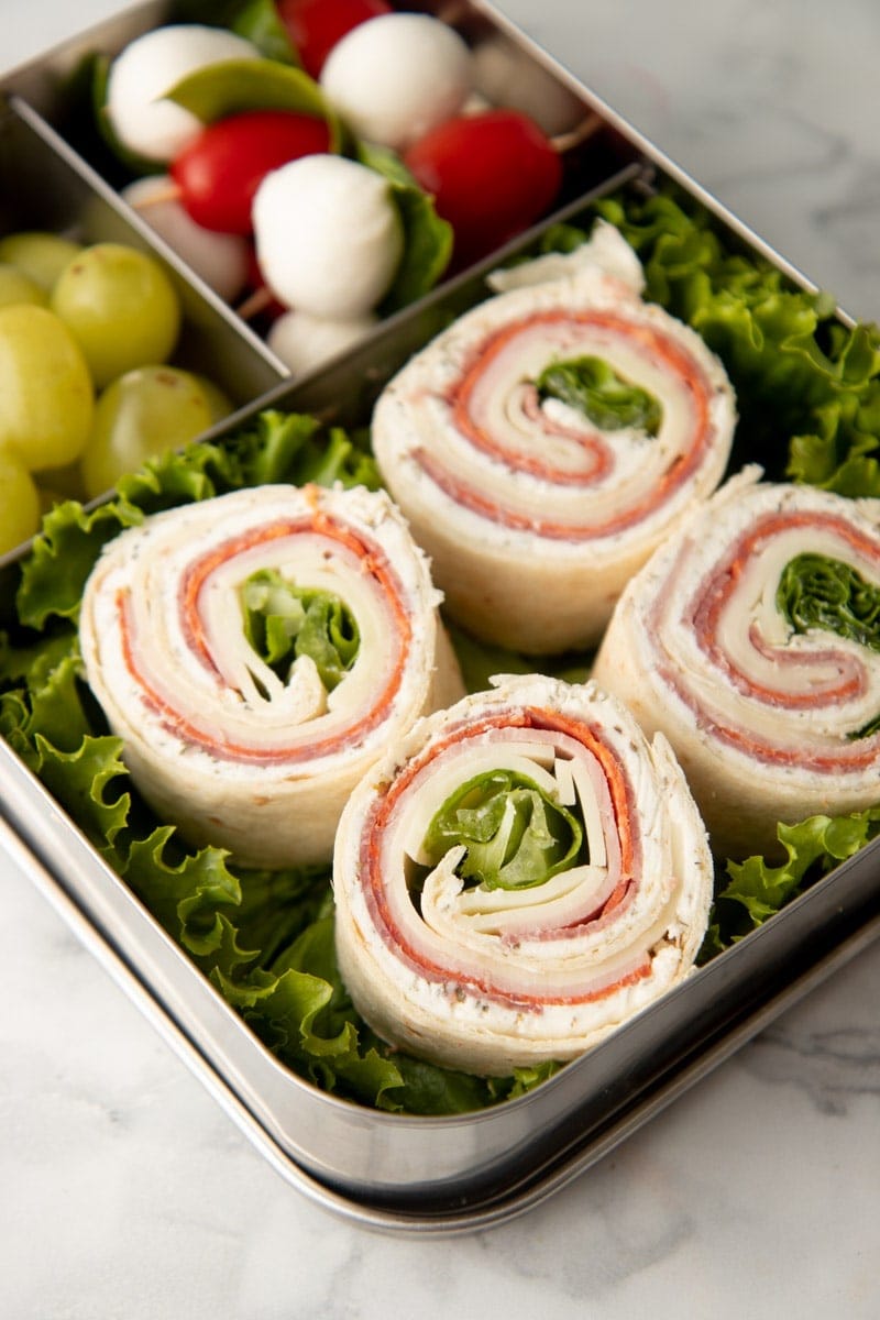 Four italian pinwheel bites nestled in a metal bento-style lunch box with green grapes and caprese sticks.