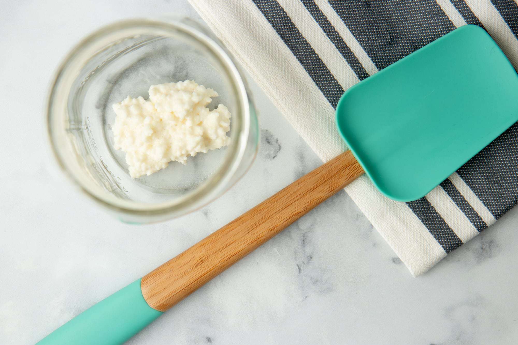 Overhead of kefir grains sitting at the bottom of a clean glass jar, a silicone scraper and linen kitchen towel sit alongside.