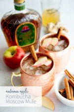 Autumn Apple Kombucha Whiskey Moscow Mule in hammered copper mugs with apple slices and a cinnamon stick floating on top, with the ingredients for the drink behind the cups. Text overlay lists the name of the drink.