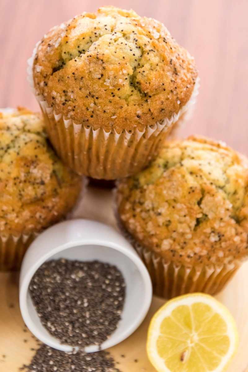 One lemon poppyseed muffin stacked atop two others, a small overturned bowl of additional seeds spilling out in front of them.