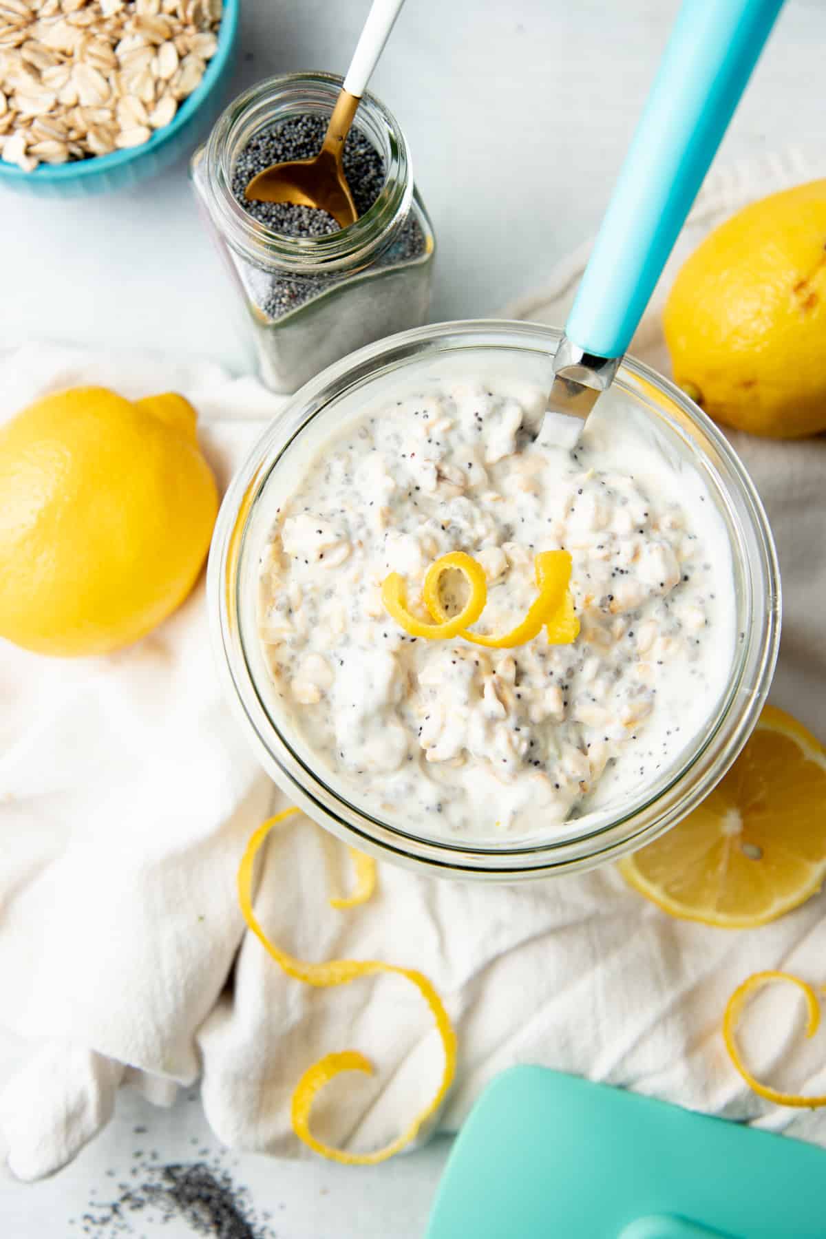 A spoon with a blue handle sits in a jar of overnight oats. Lemons and lemon zest rest on a white counter beside the jar.