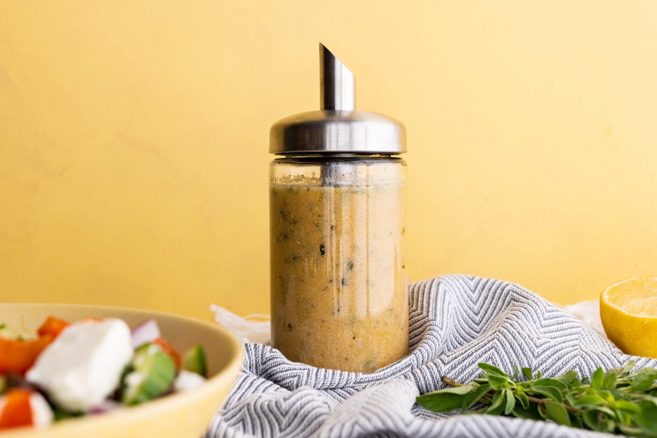 A glass cruet filled with a simple homemade lemon vinaigrette stands on a kitchen linen alongside a fresh salad.