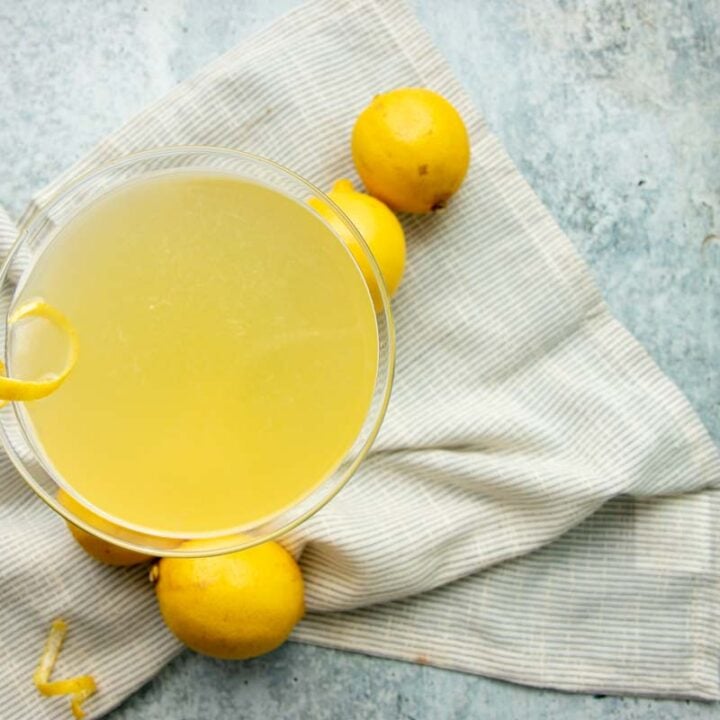 Top view of a limoncello cocktail on a cloth napkin with lemons around it on a grey counter.