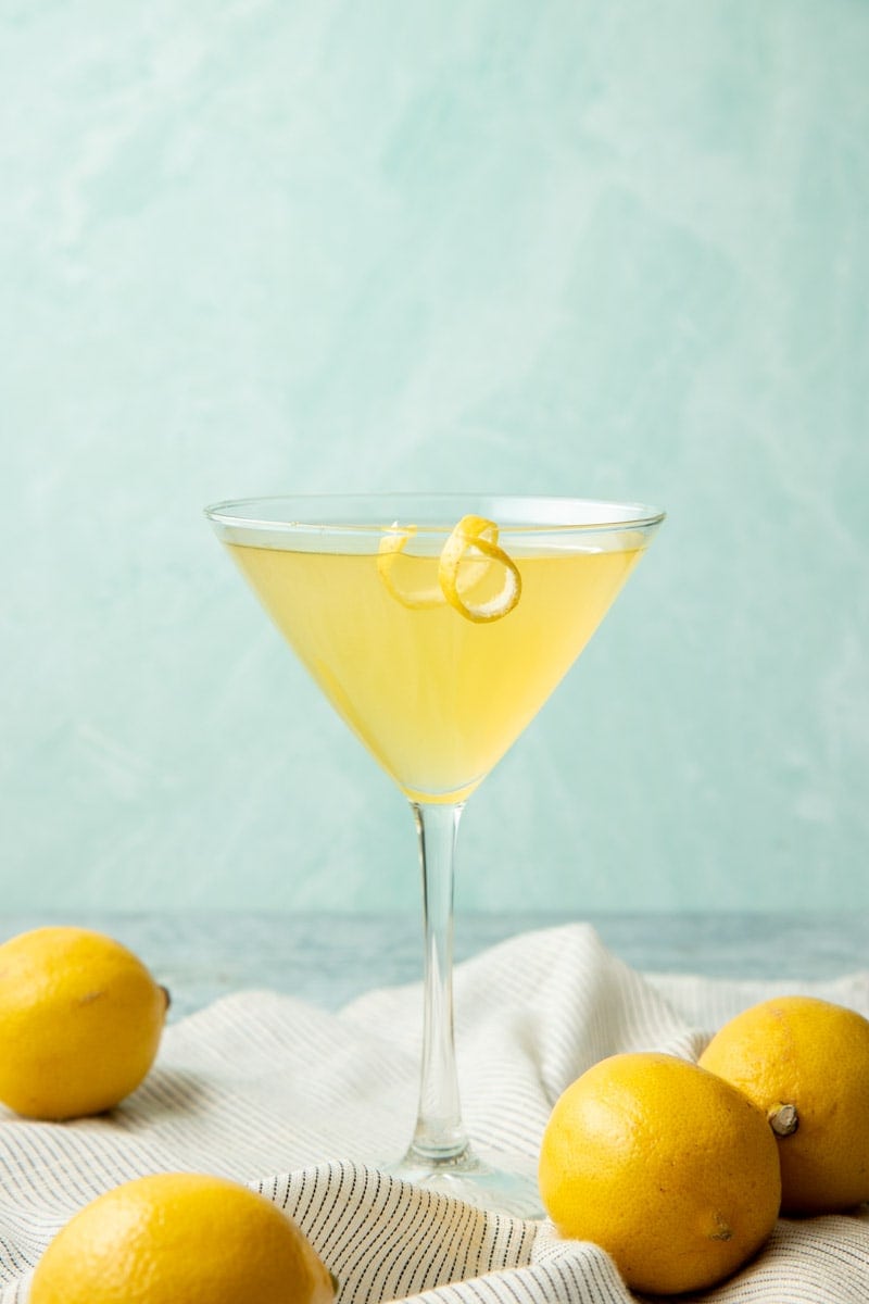 Front view of a limoncello martini in a martini glass with a lemon twist on the rim, whole lemons near the foot of the glass.