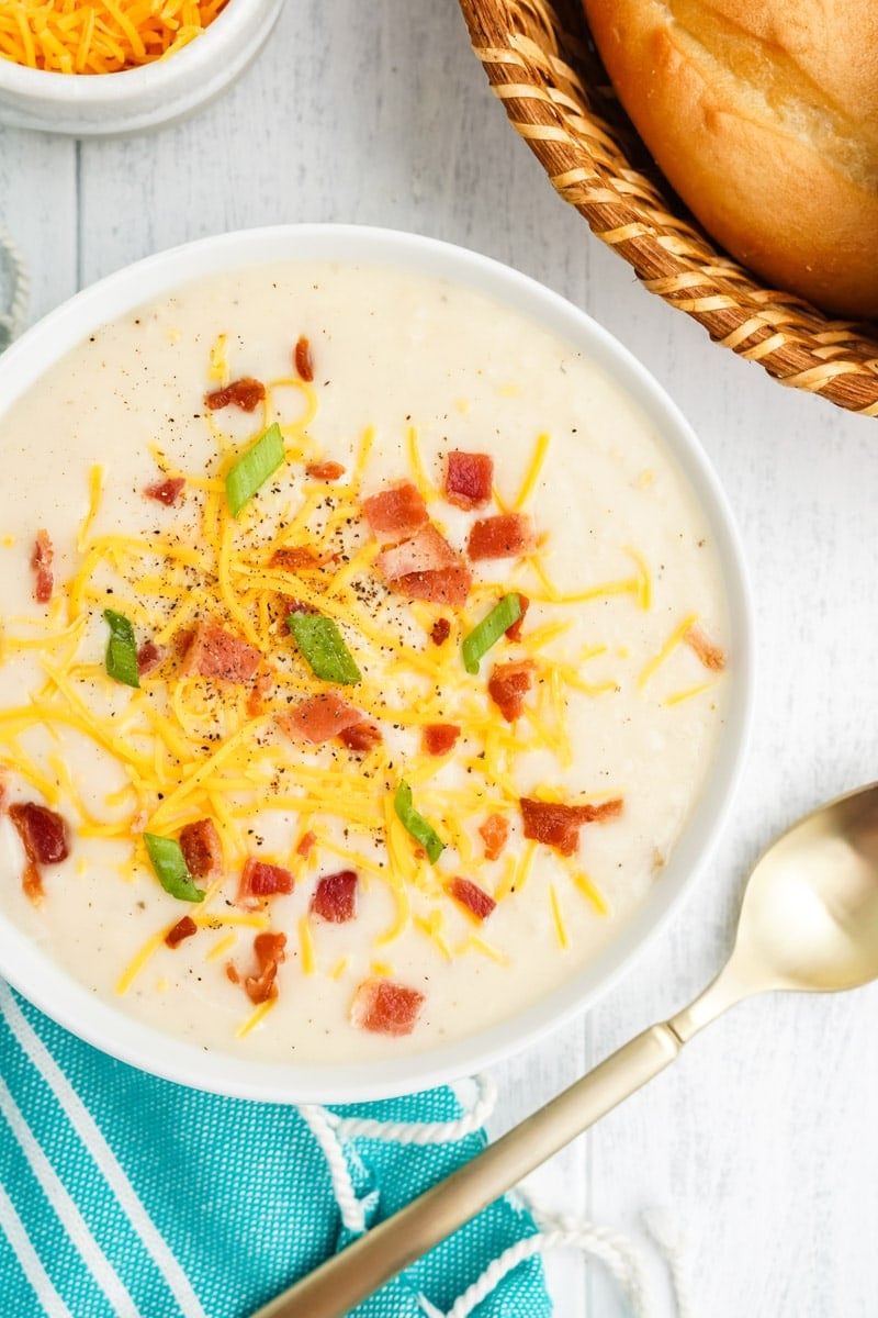 Overhead of a bowl of loaded potato soup with bread and shredded cheese nearby.