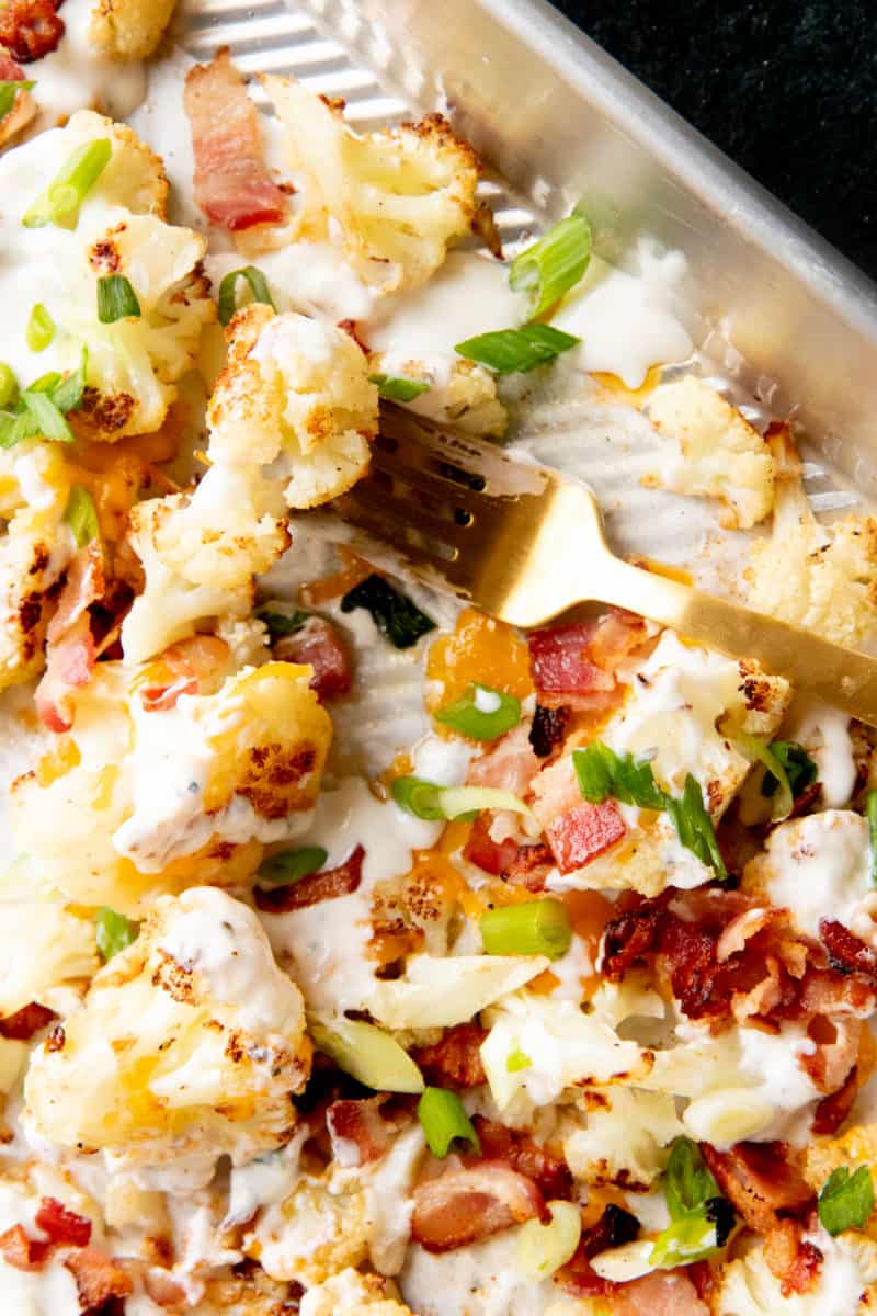 A gold fork lays on a baking pan covered in loaded roasted cauliflower.