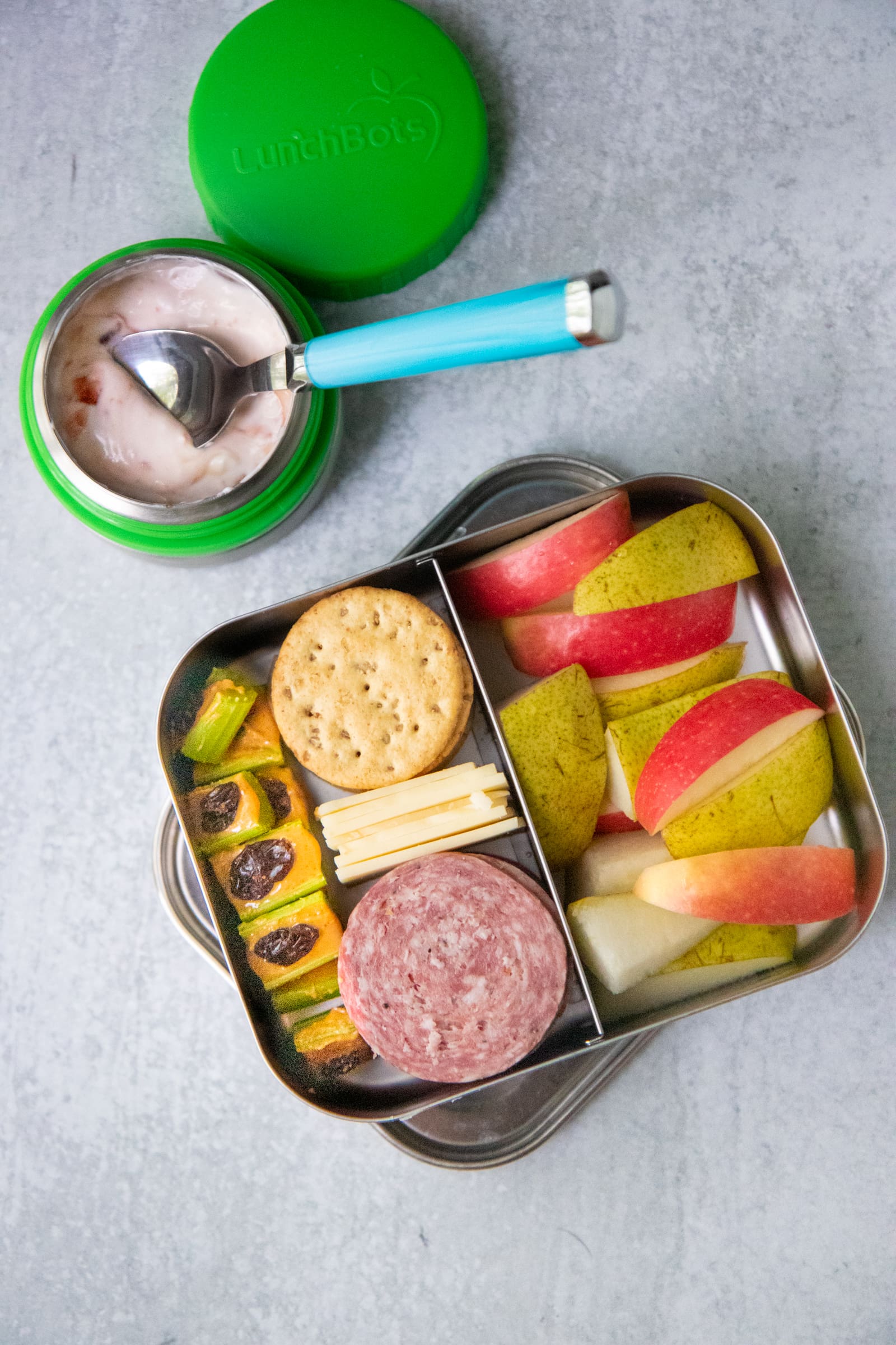 Divided lunch container filled with cheese, crackers, salami, fruit slices, and ants on a log. There is a container of yogurt next to it.