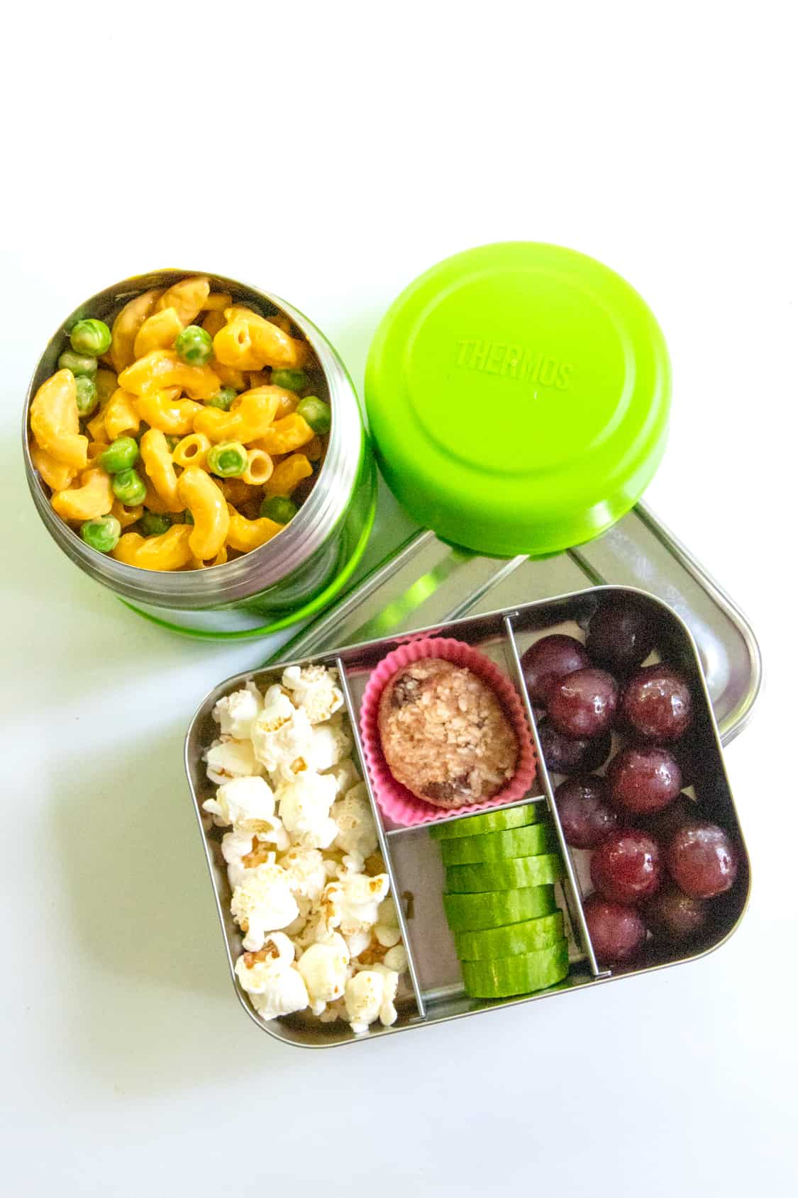 School lunch idea: Bento-style lunch box with popcorn, grapes, cucumber, and cookies next to a Thermos of mac and cheese. 