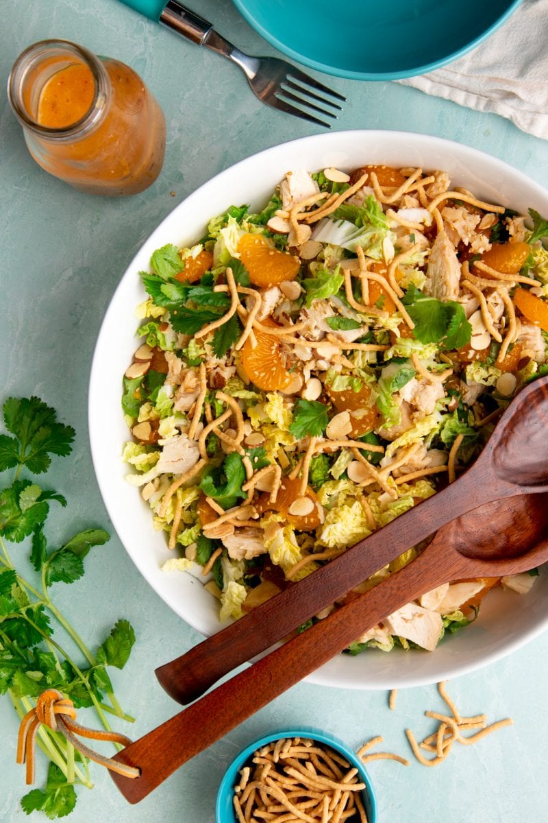 Overhead of large serving bowl of finished chicken salad and arranged around it are the homemade dressing, fresh cilantro, and crunchy chow mein noodles to garnish.