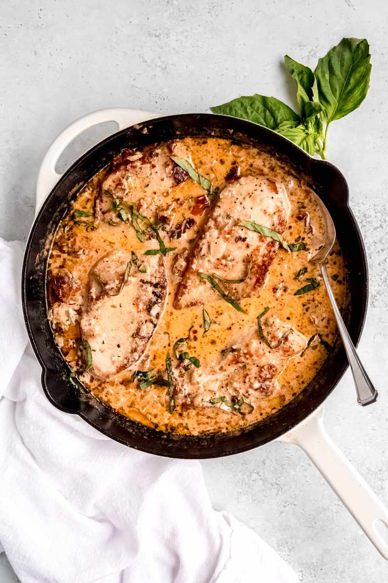 A skillet of marry me chicken garnished with fresh basil rests on a light countertop with a sprig of fresh basil tucked beside it.