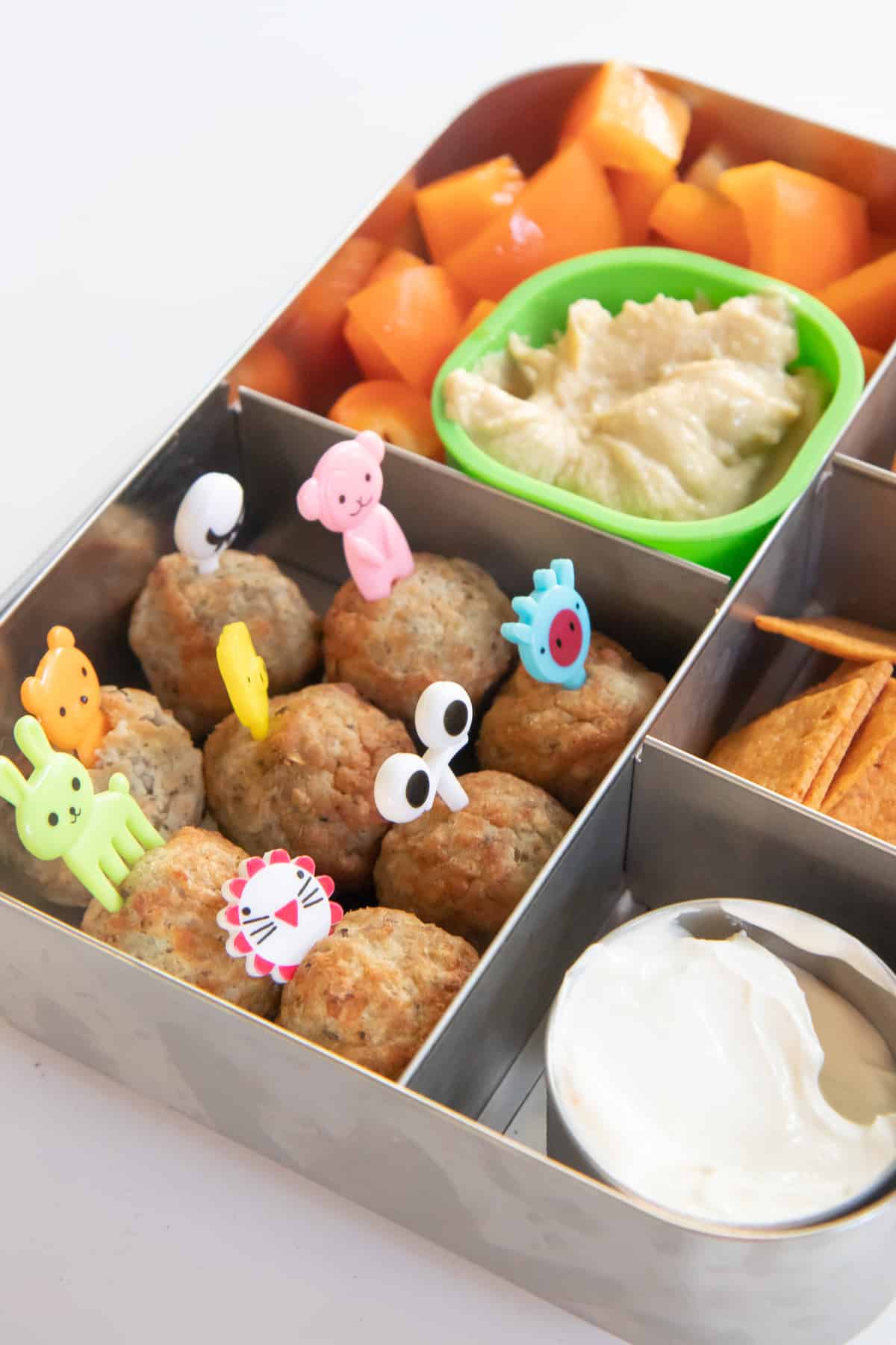 Bento-style lunchbox filled with meatballs, cantaloupe, crackers, and hummus