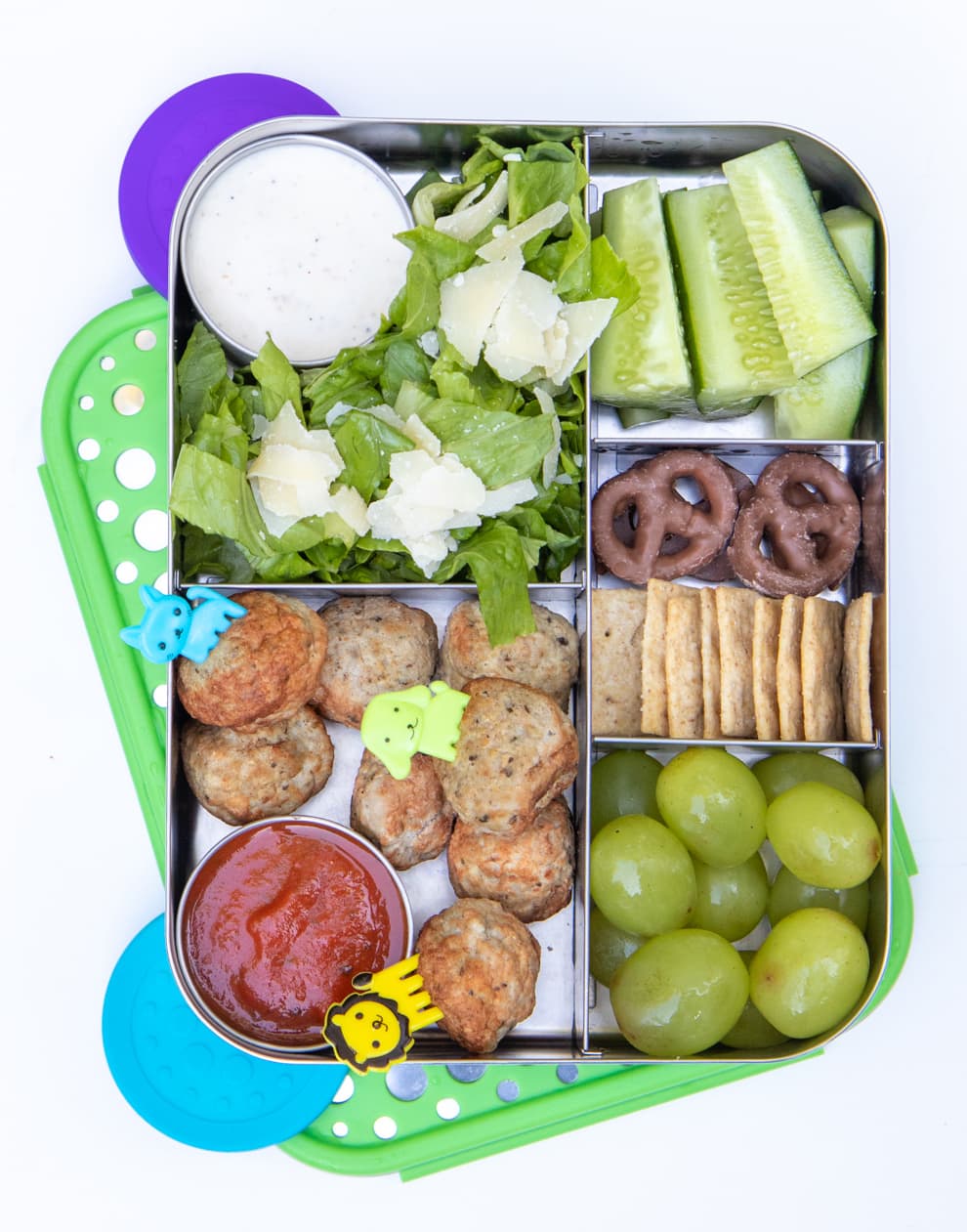 Stainless steel divided lunch box filled with meatballs, grapes, celery, pretzels, salad, and crackers.