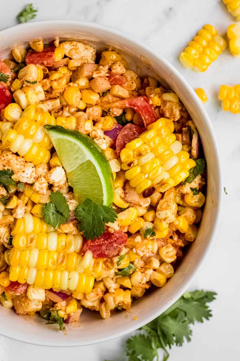 Tight view of a large white serving bowl of creamy corn salad garnished with cilantro and a lime wedge.