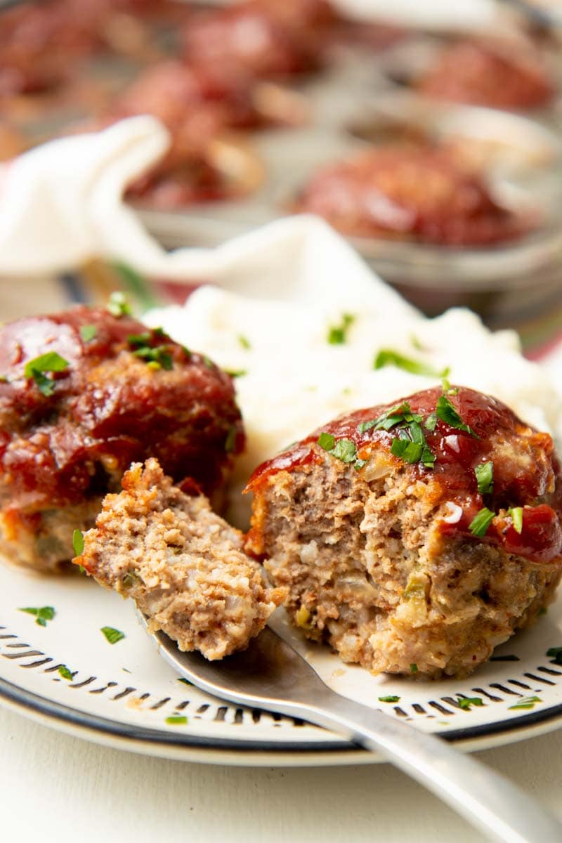 A bite of mini meatloaf rests on a fork alongside two mini meatloaves.