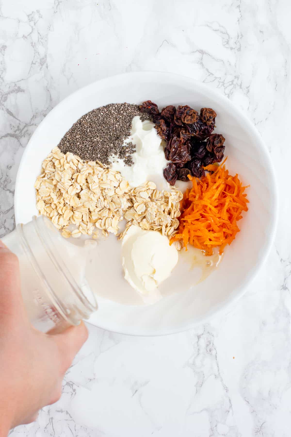 A hand pours milk from a glass jar into a white bowl. The white bowl is filled with other ingredients, including cream cheese, yogurt, shredded carrots, raisins, chia seeds, and rolled oats.