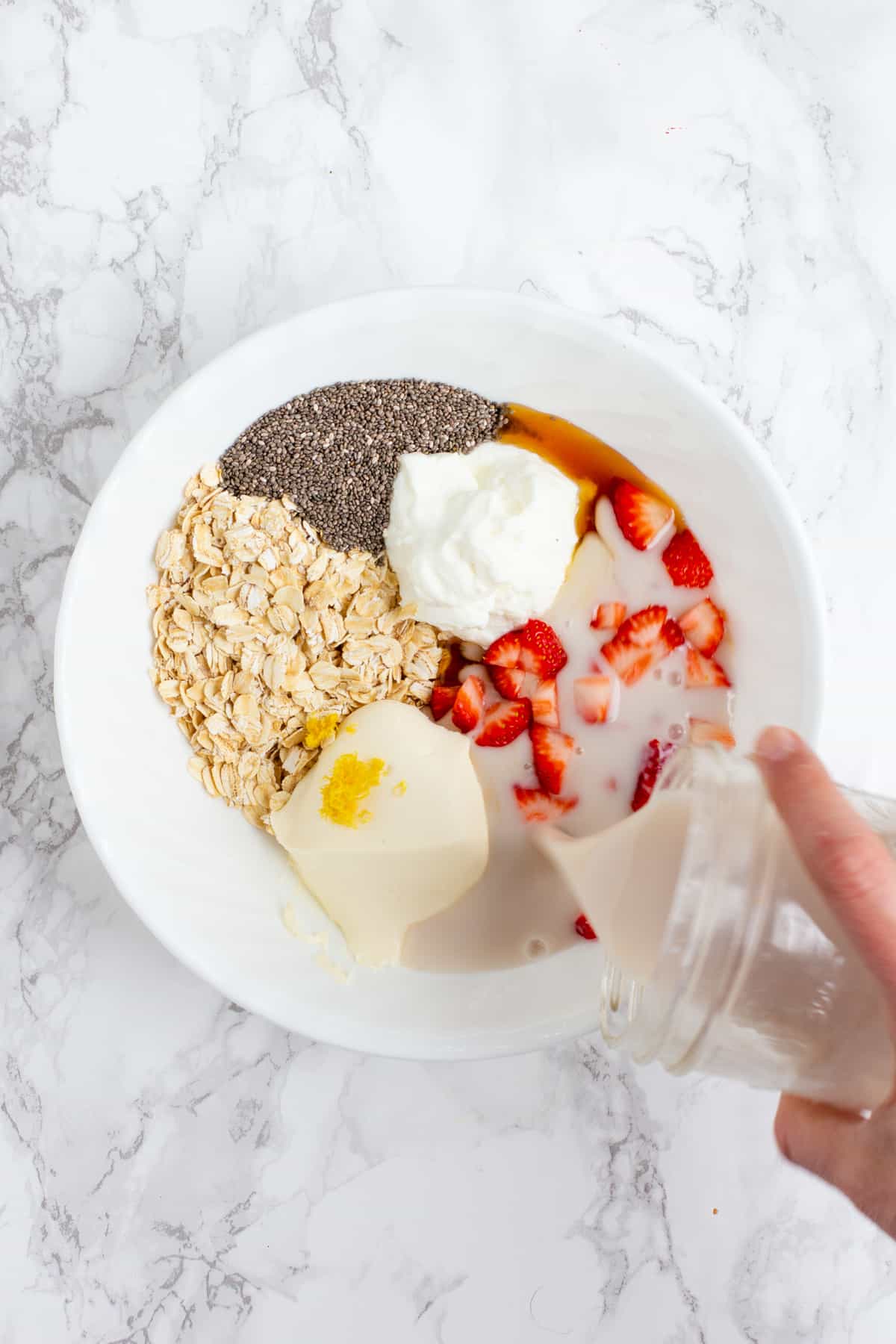 A hand pours milk from a glass jar into a white bowl. The white bowl is filled with other ingredients, including yogurt, strawberries, cream cheese, chia seeds, and rolled oats.