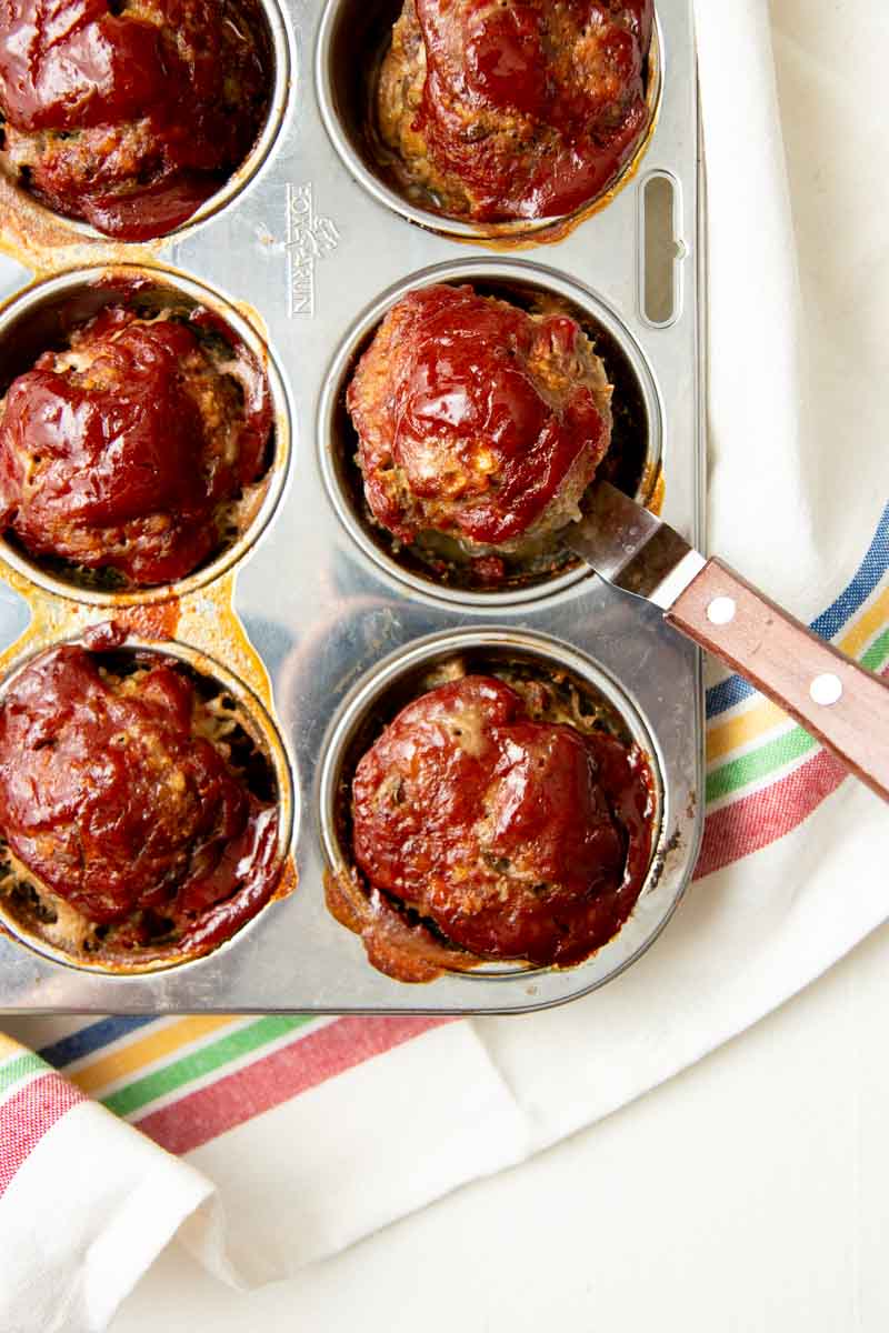 An offset spatula lifts an individual meatloaf out of a muffin tin.