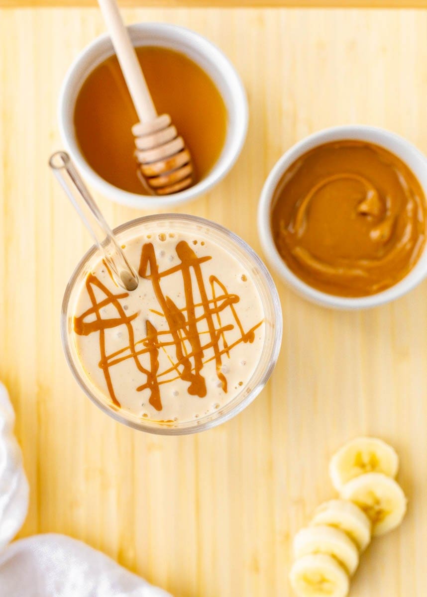 Top down view of peanut butter drizzle on top of banana smoothie with honey, banana slices, and peanut butter alongside.